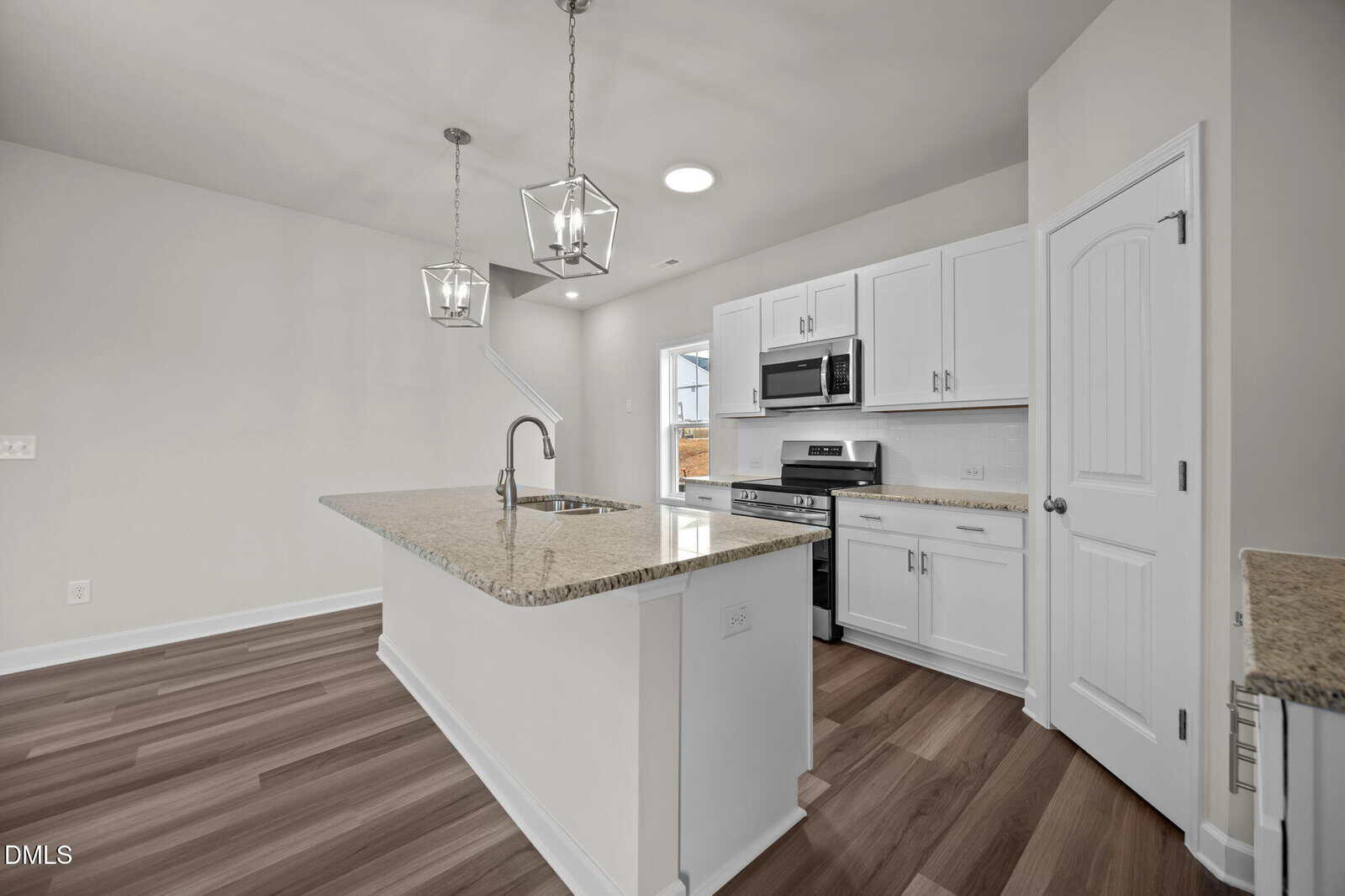 Modern kitchen with white cabinets, granite countertops, stainless steel appliances, a center island with sink, pendant lights, and wood-look flooring. A window and door provide natural light in the background.