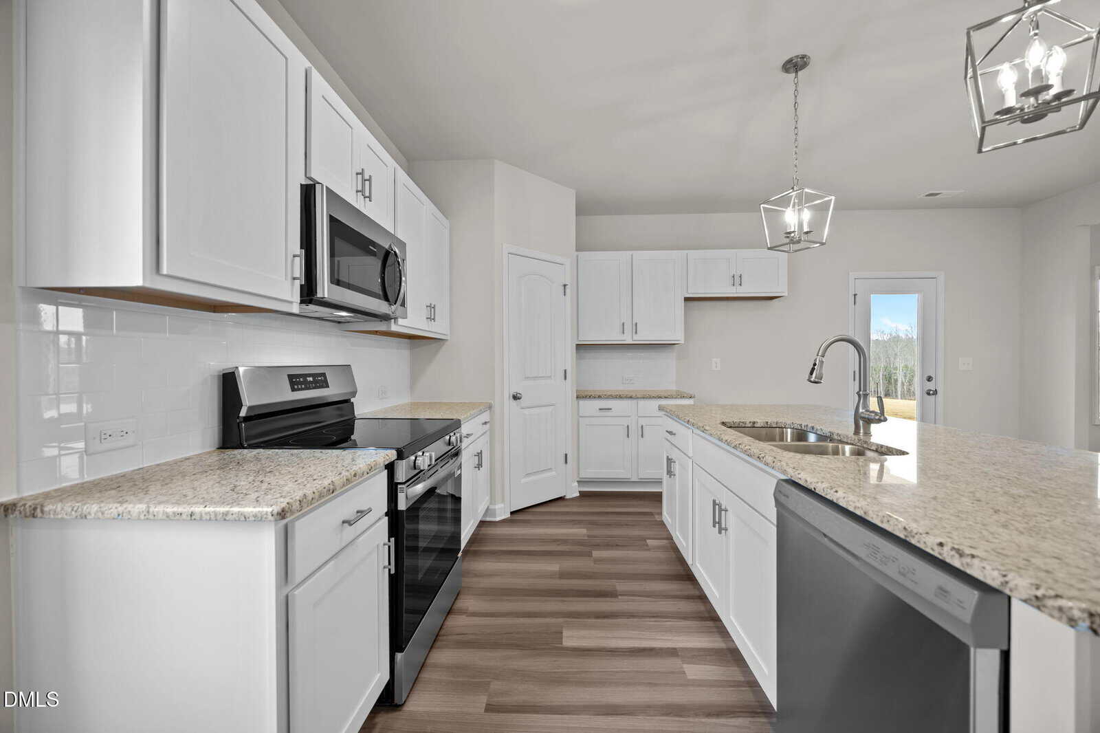 Modern kitchen with white cabinets, granite countertops, stainless steel appliances, and light wood flooring. Two pendant lights hang over a large island with a sink and dishwasher. Natural light enters through a glass door in the background.