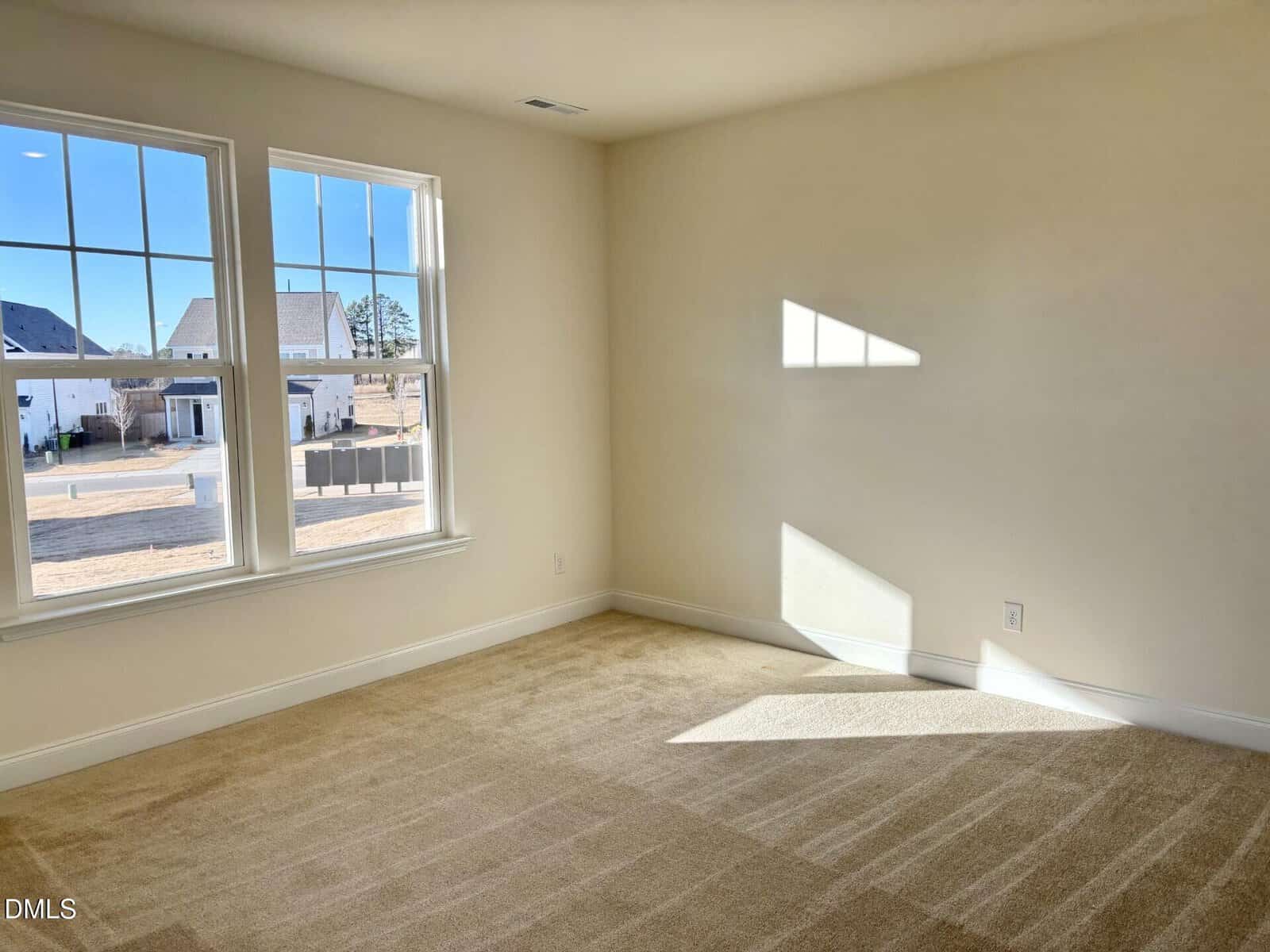 A bright, empty room with beige carpet, cream-colored walls, and two large windows letting in sunlight. Outside, a suburban neighborhood with houses and a blue sky is visible. Shadows fall on the floor and wall.