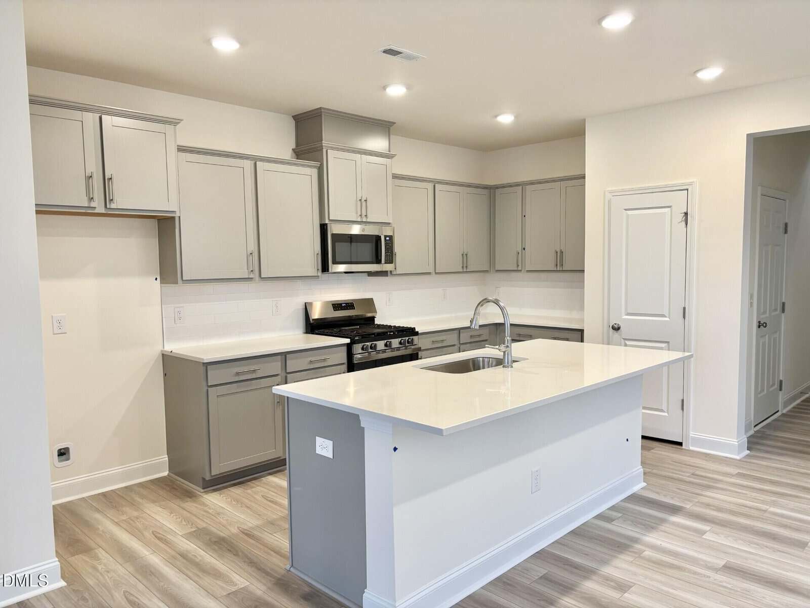 Modern kitchen with light gray cabinets, stainless steel appliances, a large white island with sink, light wood flooring, white subway tile backsplash, and recessed ceiling lights.