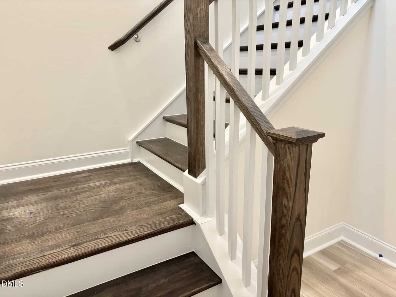 A close-up view of a modern indoor staircase with dark wooden steps and white risers, featuring a wooden handrail and white balusters. The walls are light-colored, and the flooring at the base is a light wood.