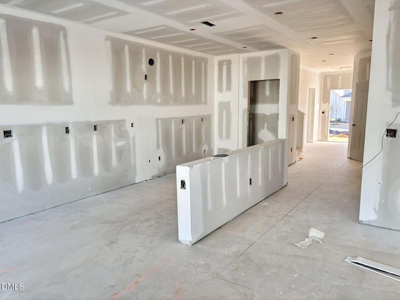 A room under construction with unfinished drywall, exposed electrical outlets, and no flooring or fixtures. Sunlight enters through an open door at the far end, and construction materials are scattered on the concrete floor.