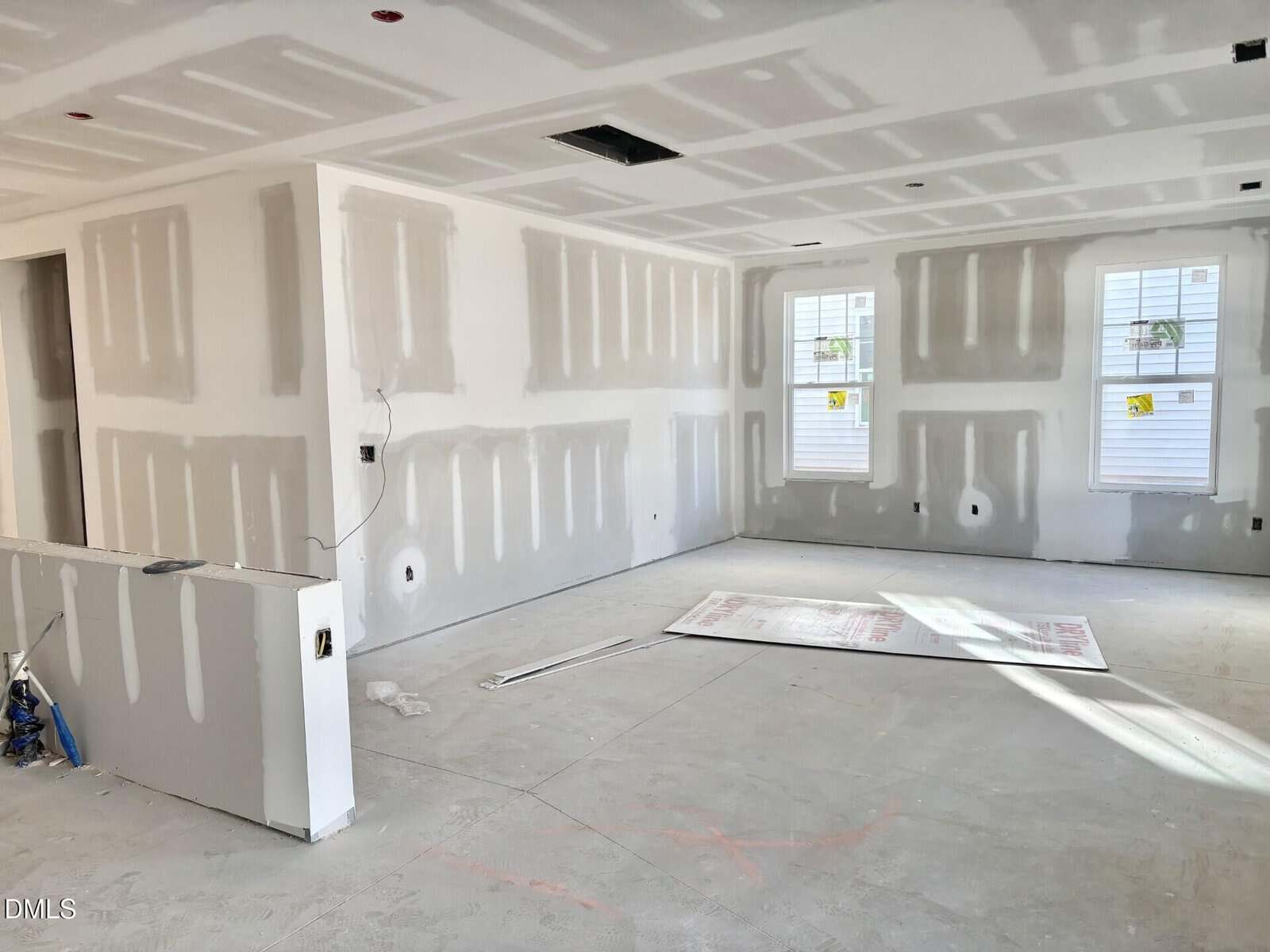 A room under construction with unfinished drywall, exposed seams, two windows, and construction materials on the floor. The space is bright with natural light coming through the windows.