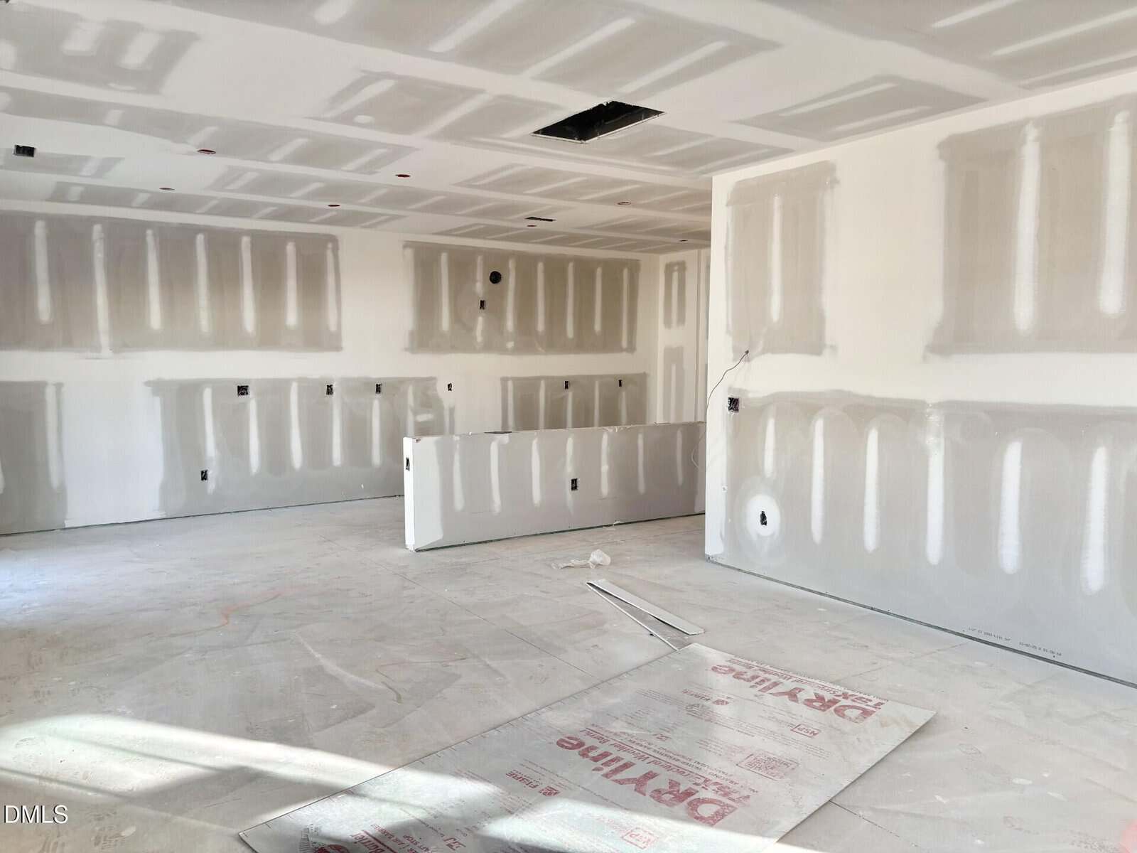 A room under construction with unfinished drywall, exposed joints, and no flooring installed. Tools and materials are scattered on the floor, and light enters from the left, illuminating the space.