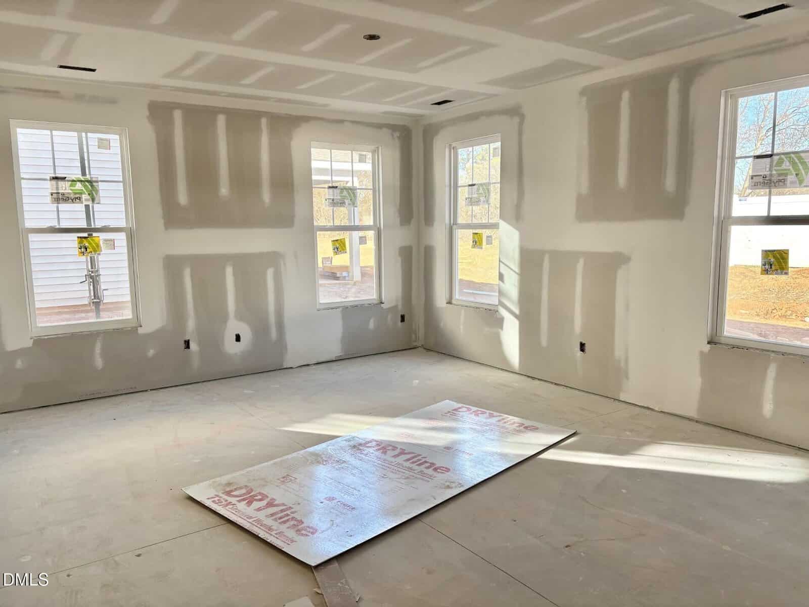 A sunlit room under construction features unpainted drywall, five large windows, and a protective sheet on the floor. Shadows and construction materials are visible.