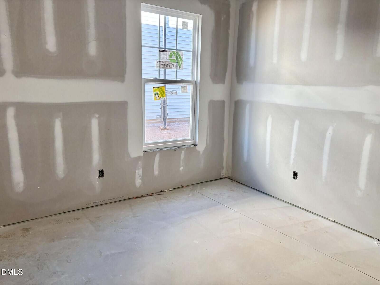 Unfinished room with drywall installed but not painted, visible joint compound lines, a single window with stickers, and an unfinished floor. Blue building seen outside through the window.