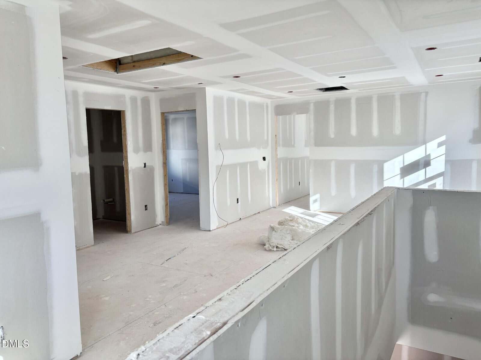 A bright, unfinished room with white drywall, exposed seams, open doorways, and no flooring. Sunlight streams through a window, casting shadows on the walls. Construction materials are scattered on the floor.