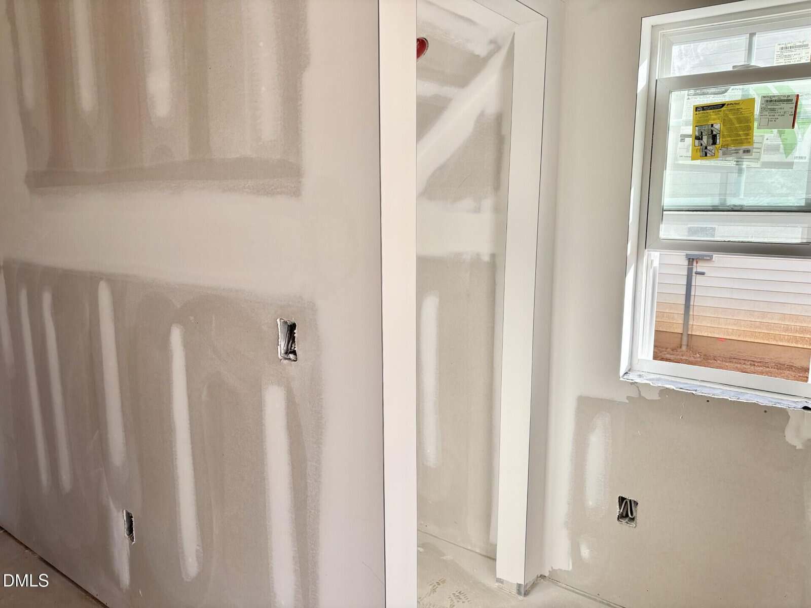 A room under construction with unfinished drywall, exposed electrical outlets, a window with a building permit attached, and natural light coming through.