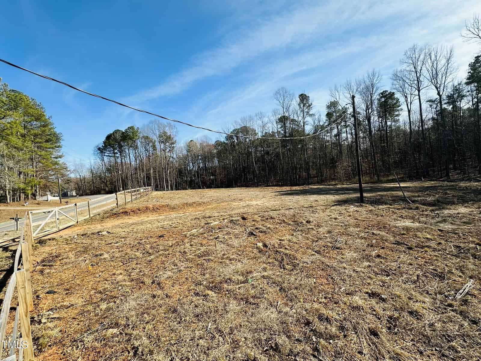 A cleared, grassy lot bordered by a wooden fence runs alongside 105 Old John Mitchell Road in Youngsville. Leafless trees line the background beneath a blue sky, and utility lines cross the scene overhead.