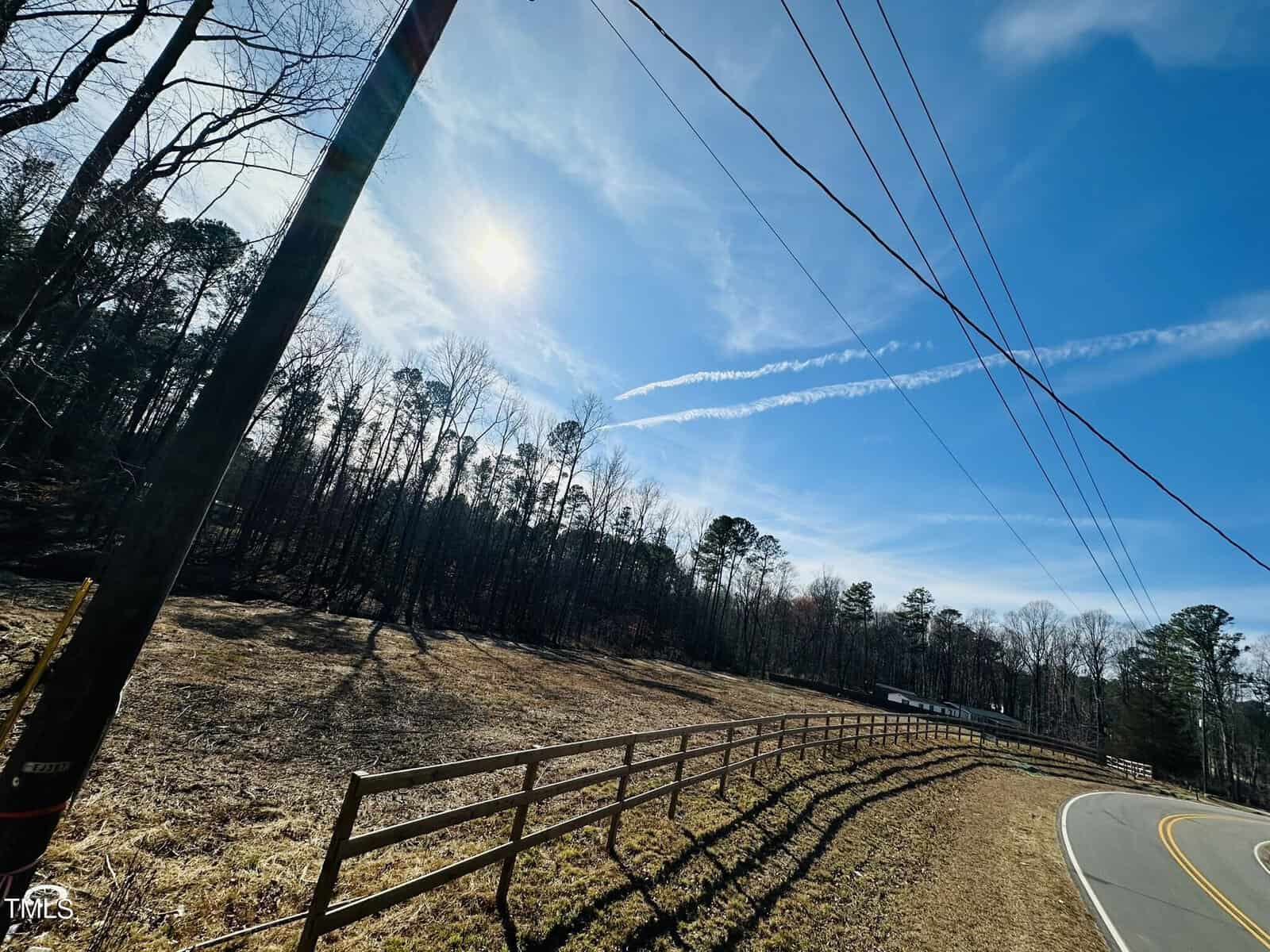 A curved rural road runs alongside a wooden fence and a field bordered by tall trees. Power lines cross the blue sky, and the sun shines brightly, casting long shadows. A white contrail appears diagonally in the sky.