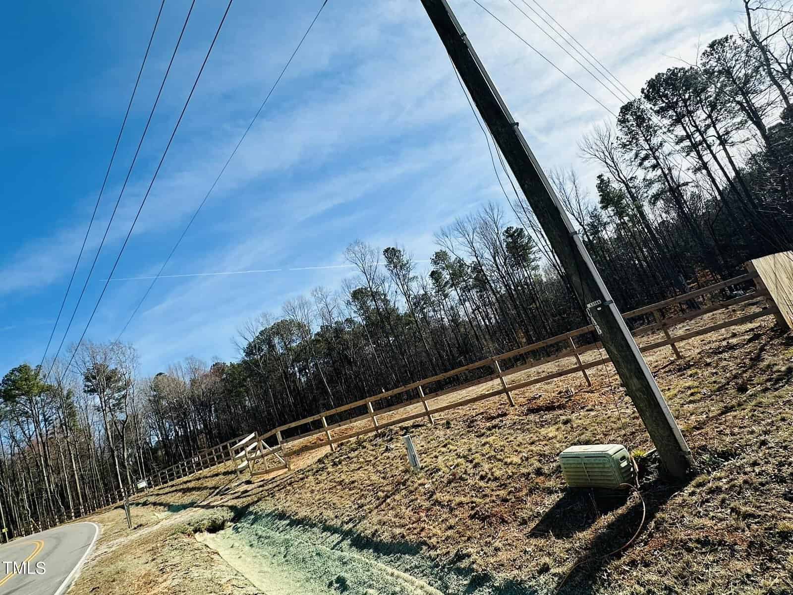 A two-lane road curves beside a fenced area with bare trees. Utility poles and wires line the road, and a green utility box is on the grassy slope. The sky is clear with a few thin clouds.