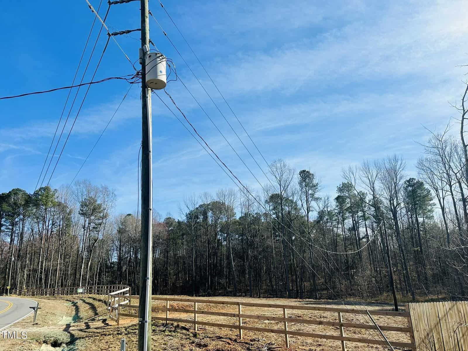 A tall utility pole with power lines stands near a wooden fence and a curved road. Leafless trees line the background under a clear blue sky. Grass and bare earth are visible in the foreground.