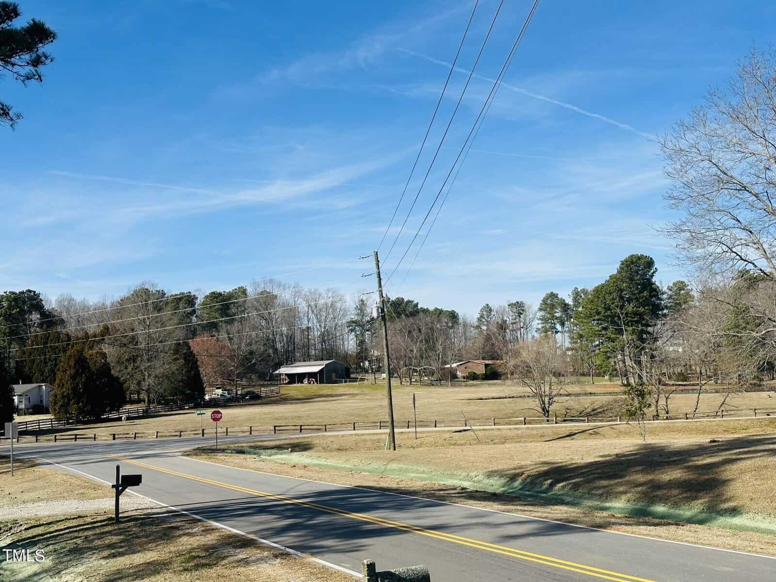 A rural road runs through a grassy area with a mailbox in the foreground. Trees and houses are in the background under a clear blue sky. Power lines cross above the road.