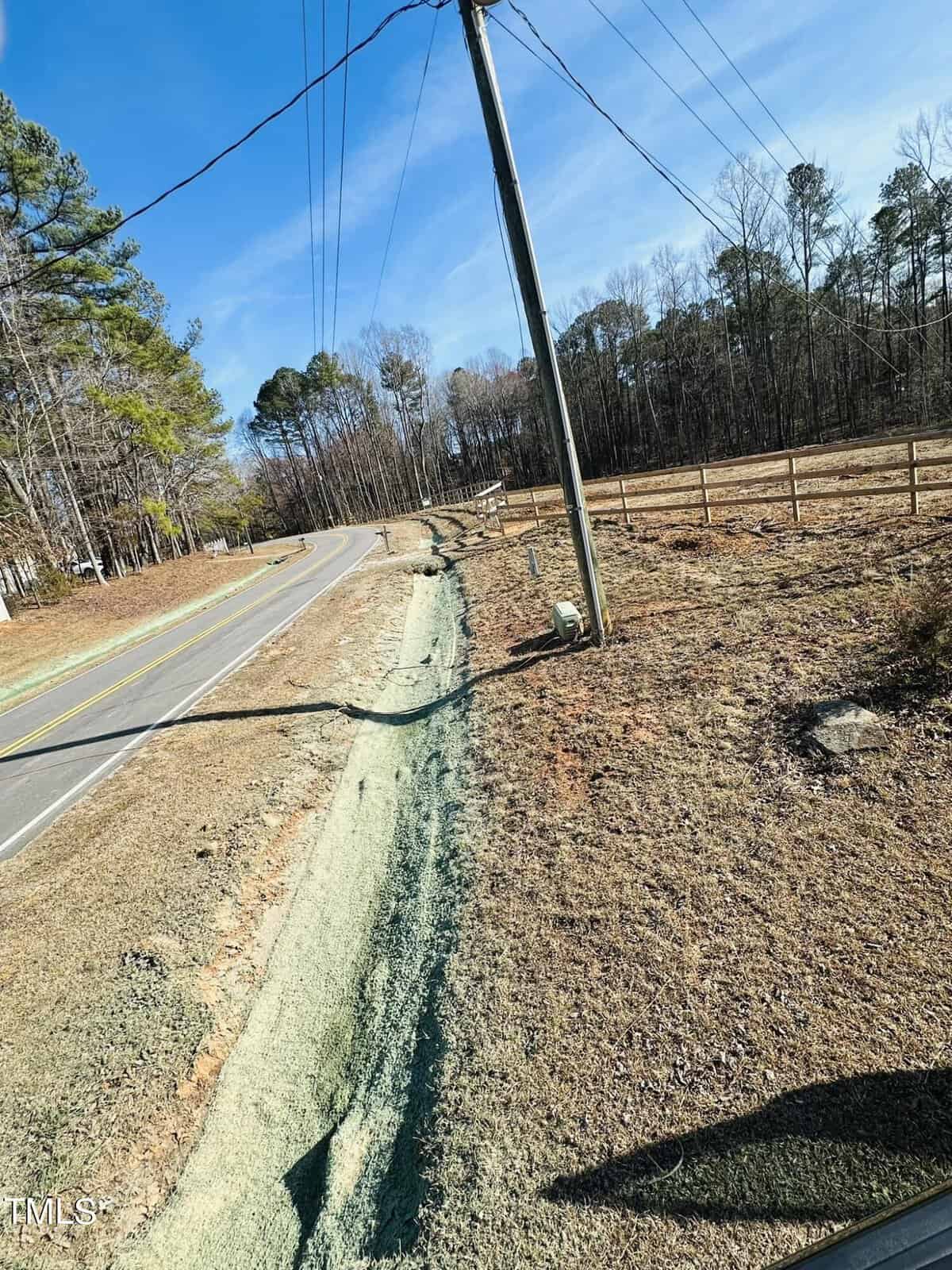 A rural roadside with a utility pole, grass seed or hydroseeding material covering a narrow strip by the road, bordered by a wooden fence and trees in the background under a clear blue sky.