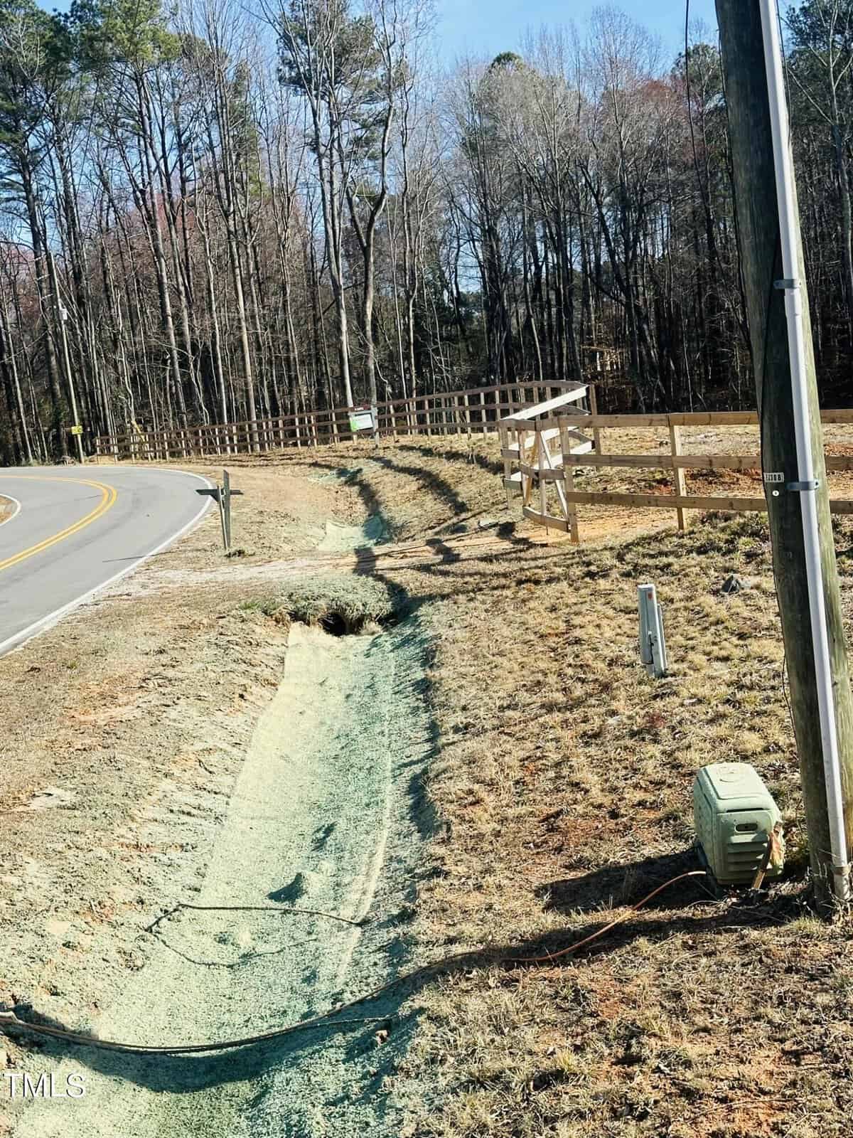 A roadside drainage ditch covered with green erosion control material runs alongside a curved road. There is a mailbox, utility boxes, a wooden fence, and trees in the background under a clear sky.