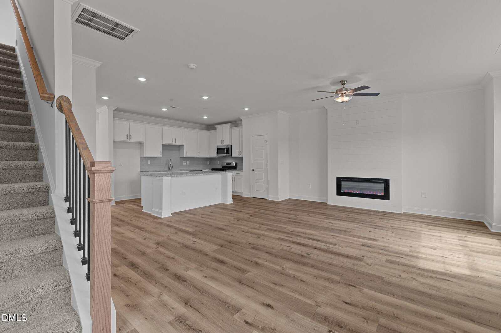 Open-plan living area with light wood flooring, white walls, a ceiling fan, an electric fireplace, and an adjacent kitchen featuring white cabinetry and a kitchen island. Carpeted stairs with a wooden handrail are on the left.