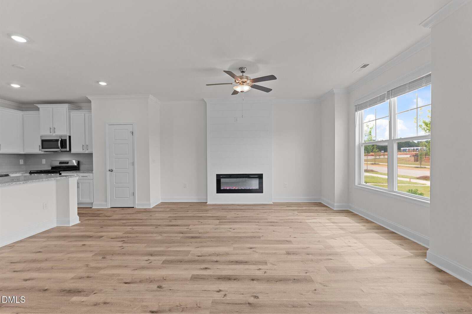 A spacious, empty living room with light wood floors, white walls, a ceiling fan, large window, and a built-in electric fireplace. The open layout connects to a white kitchen with stainless steel appliances.