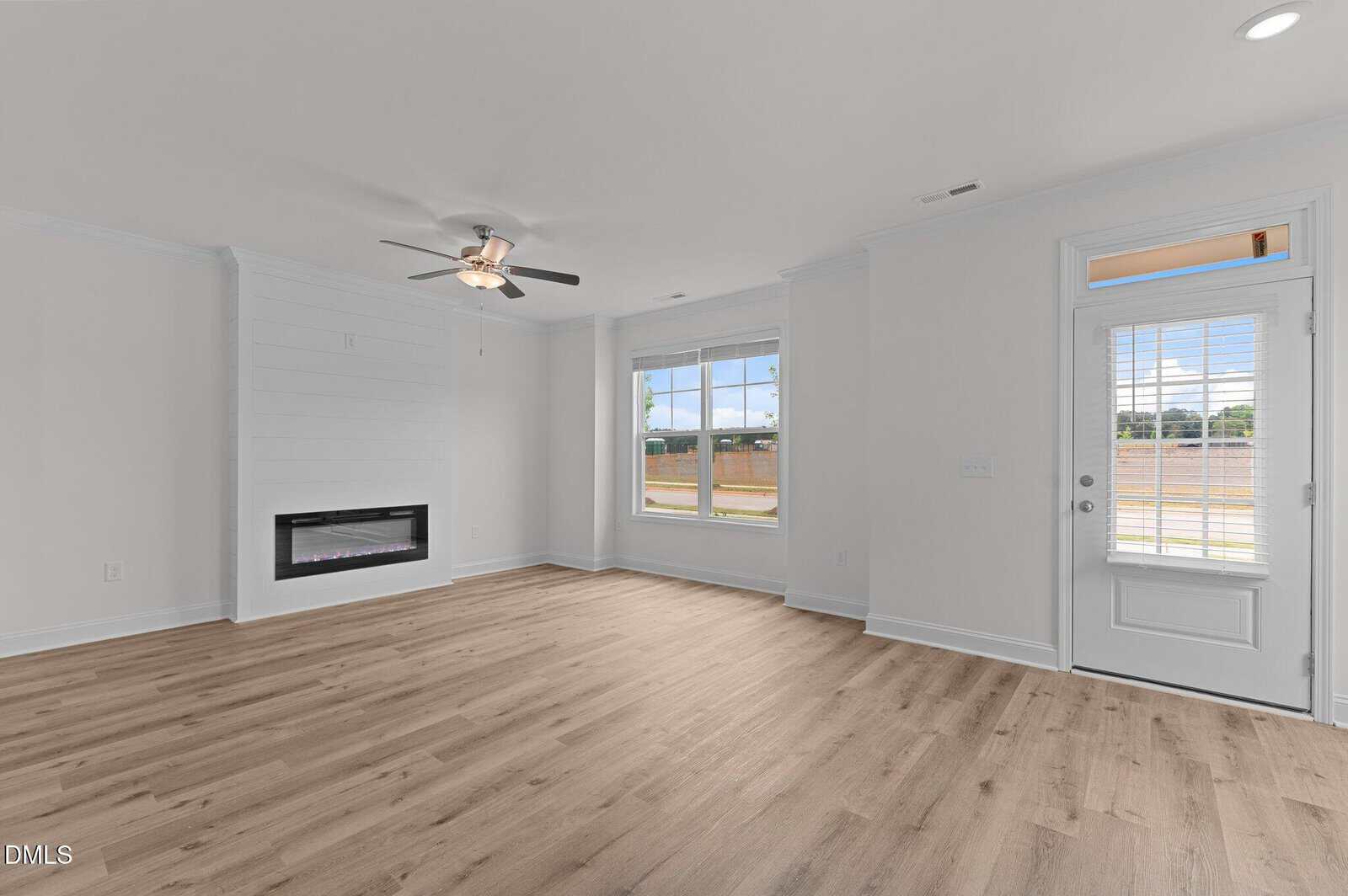 A bright, empty living room with light wood floors, white walls, a ceiling fan, large window, glass door, and a built-in modern fireplace on the left wall.