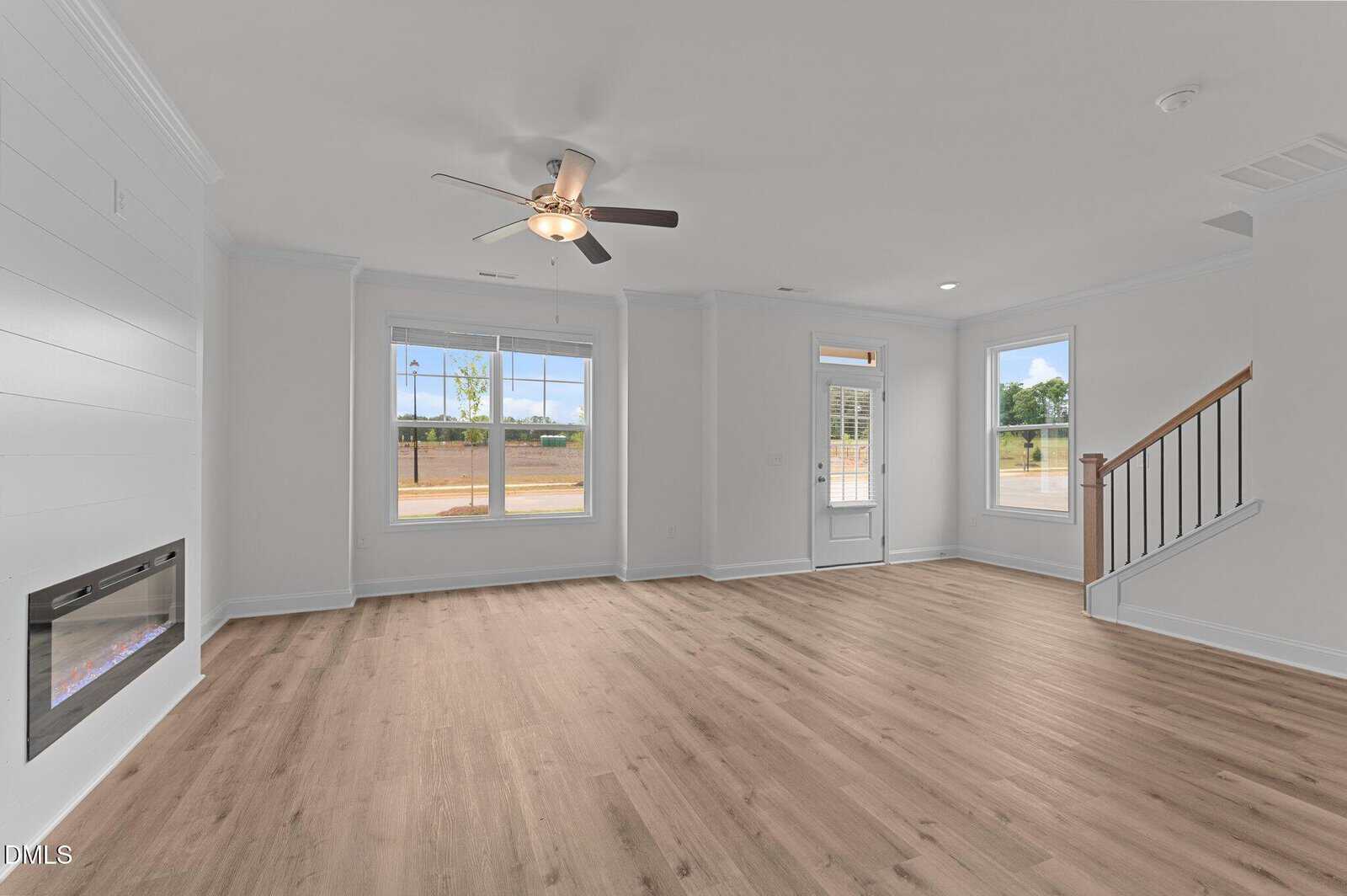 A spacious, empty living room with light wood flooring, white walls, large windows, a ceiling fan, an electric fireplace on the left wall, and a staircase on the right. Natural light fills the room.