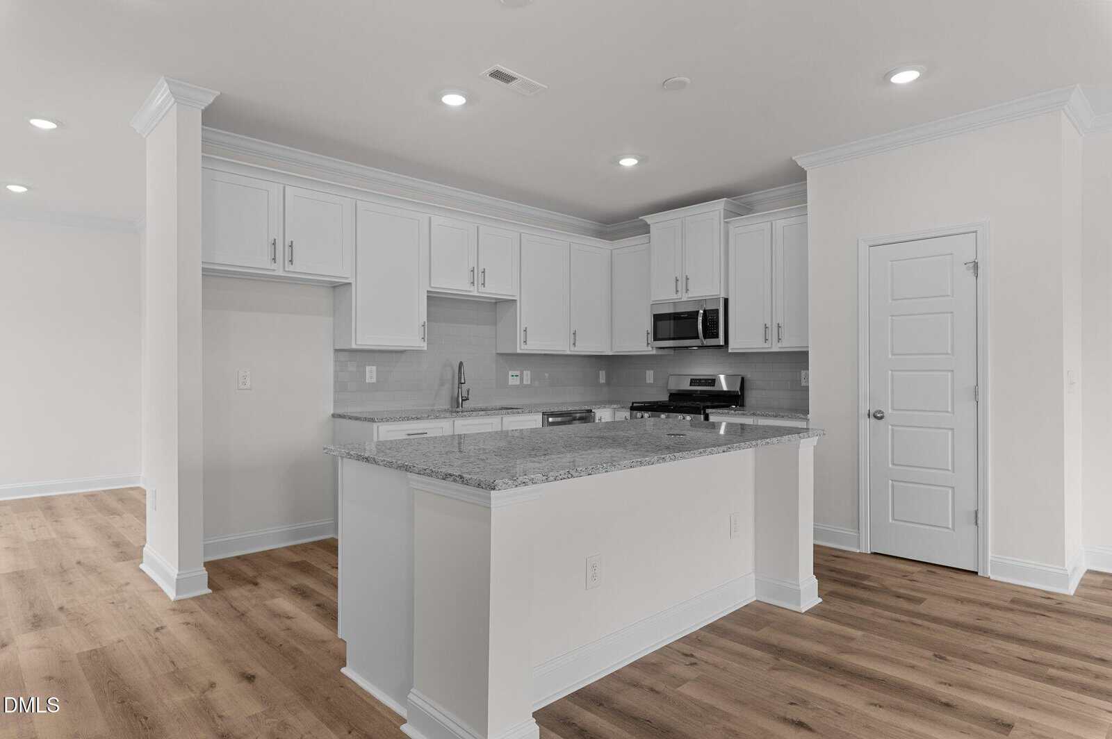 Modern kitchen with white cabinets, stainless steel appliances, a granite countertop island, light gray backsplash, and wood flooring. Recessed ceiling lights are on, and there is a closed white door to the right.