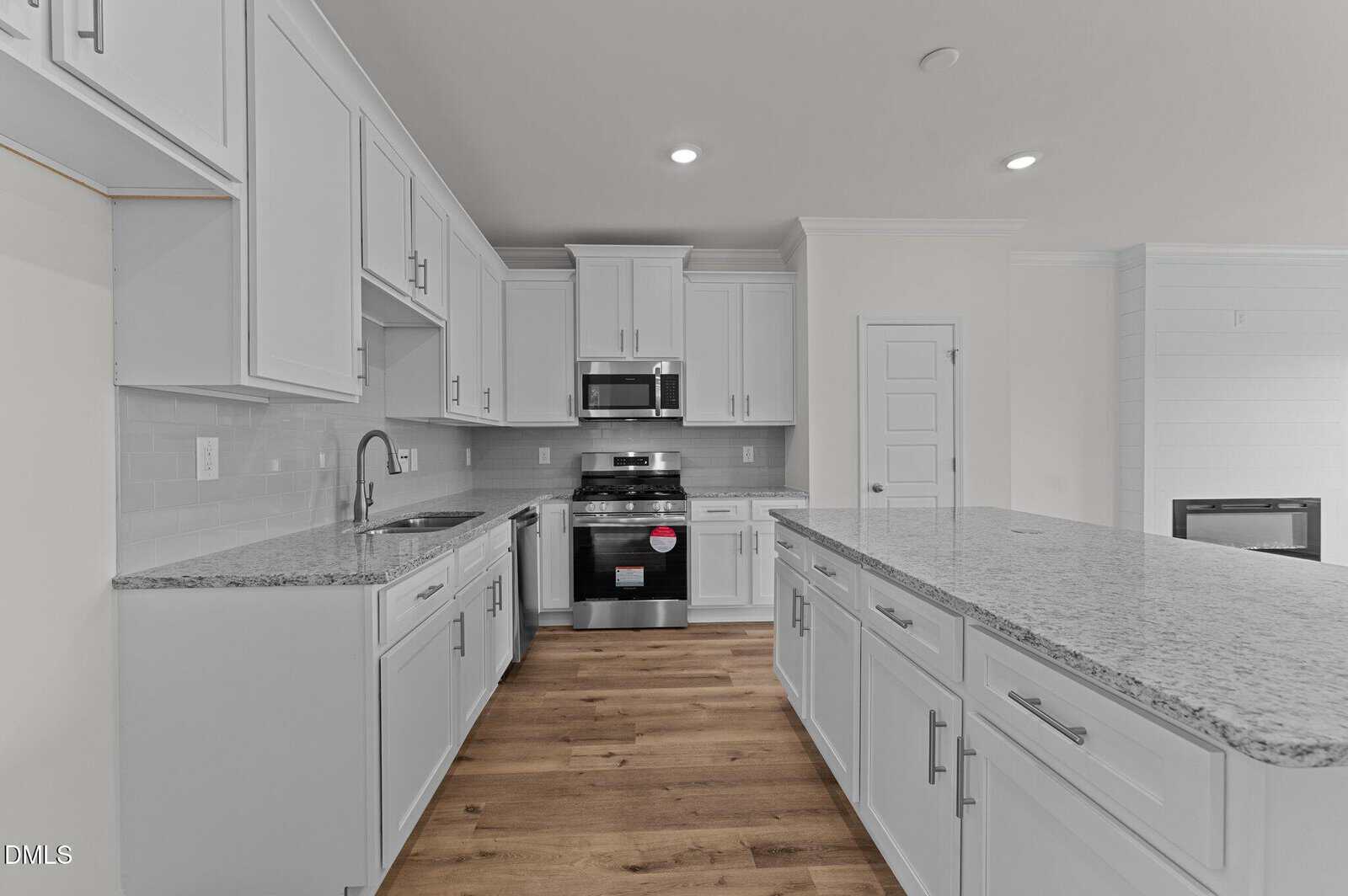 A modern kitchen with white cabinets, granite countertops, stainless steel appliances, a large island with drawers, a double sink, light gray tile backsplash, and wood flooring.