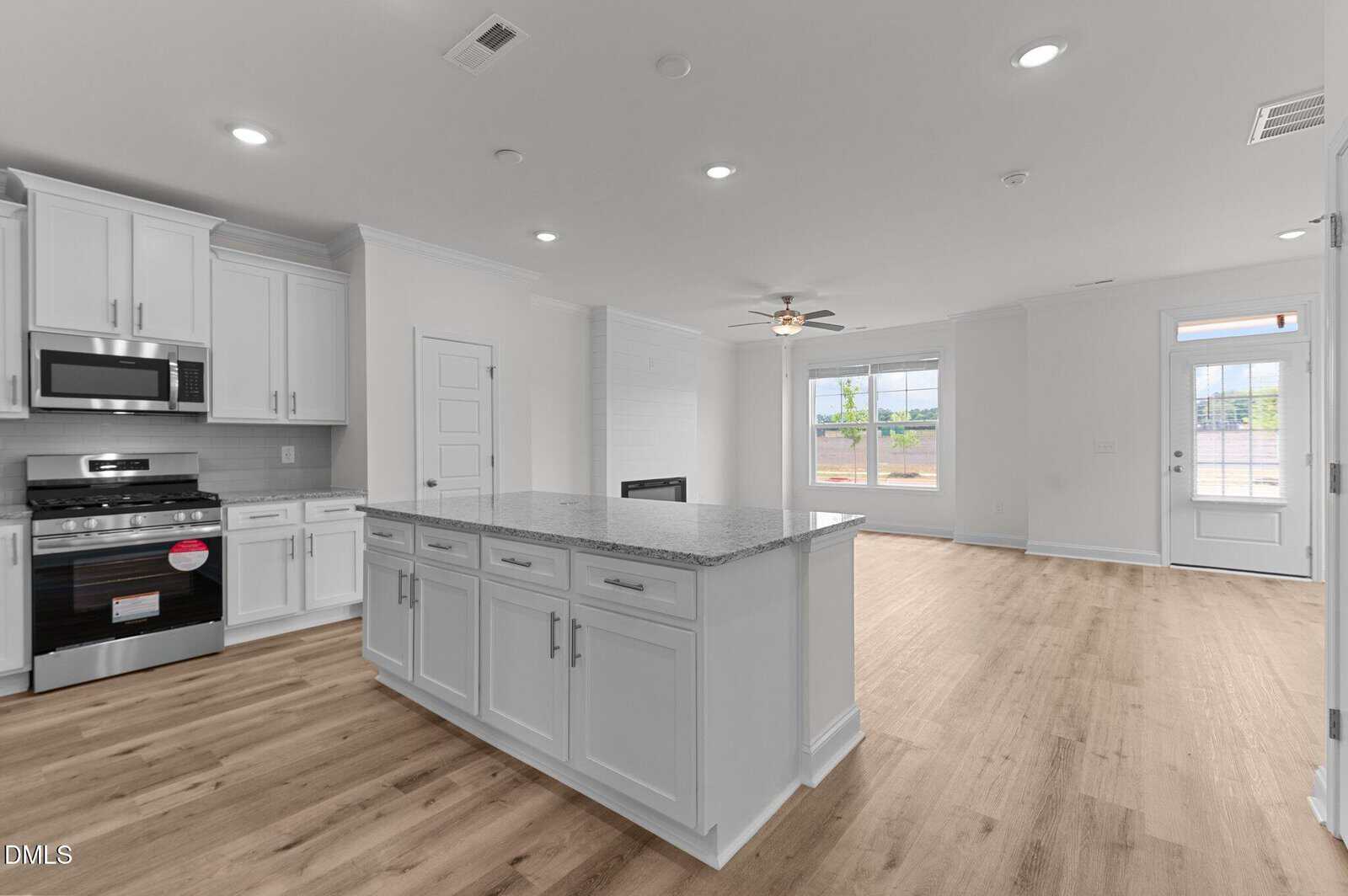 A modern open-concept kitchen with white cabinets, stainless steel appliances, a central island, and light wood flooring. The kitchen opens to a spacious living area with large windows and a ceiling fan.