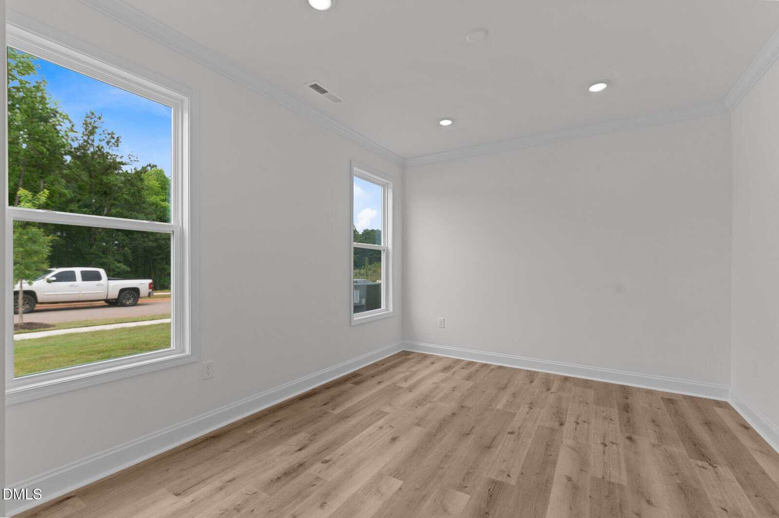 An empty room with white walls, recessed ceiling lights, two large windows letting in natural light, and light-colored wood flooring. A white truck and greenery are visible outside through the windows.