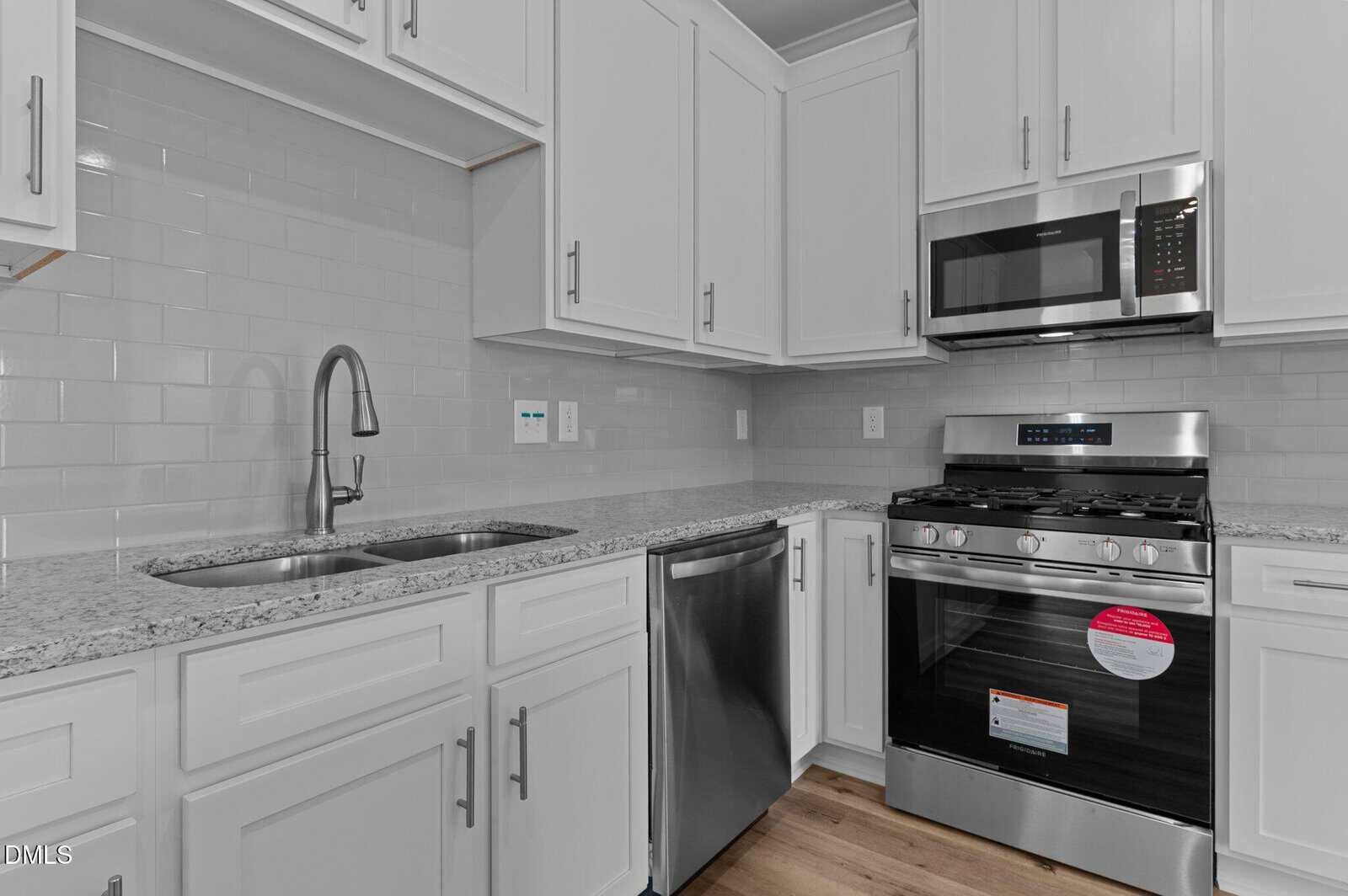 A modern kitchen with white cabinets, granite countertops, a stainless steel double sink, dishwasher, gas stove with oven, and microwave. The backsplash is white subway tile and the floor is light wood.