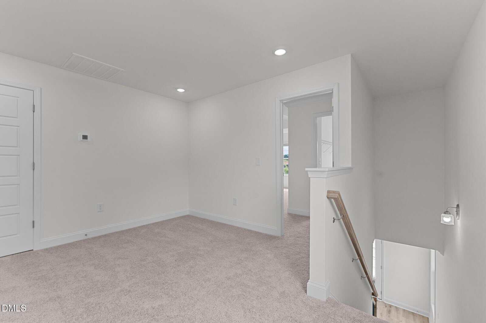 A carpeted room with white walls, a closed door, a wall control, an open doorway, and stairs leading down with a wooden handrail; recessed ceiling lights illuminate the space.