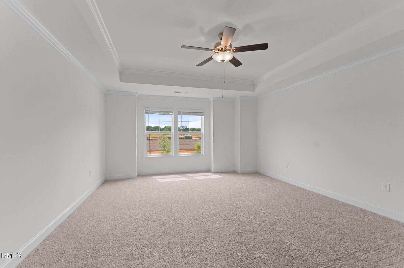 Spacious empty room with light beige carpet, white walls, a ceiling fan, and two windows letting in natural light. The view outside shows greenery and a wooden fence.