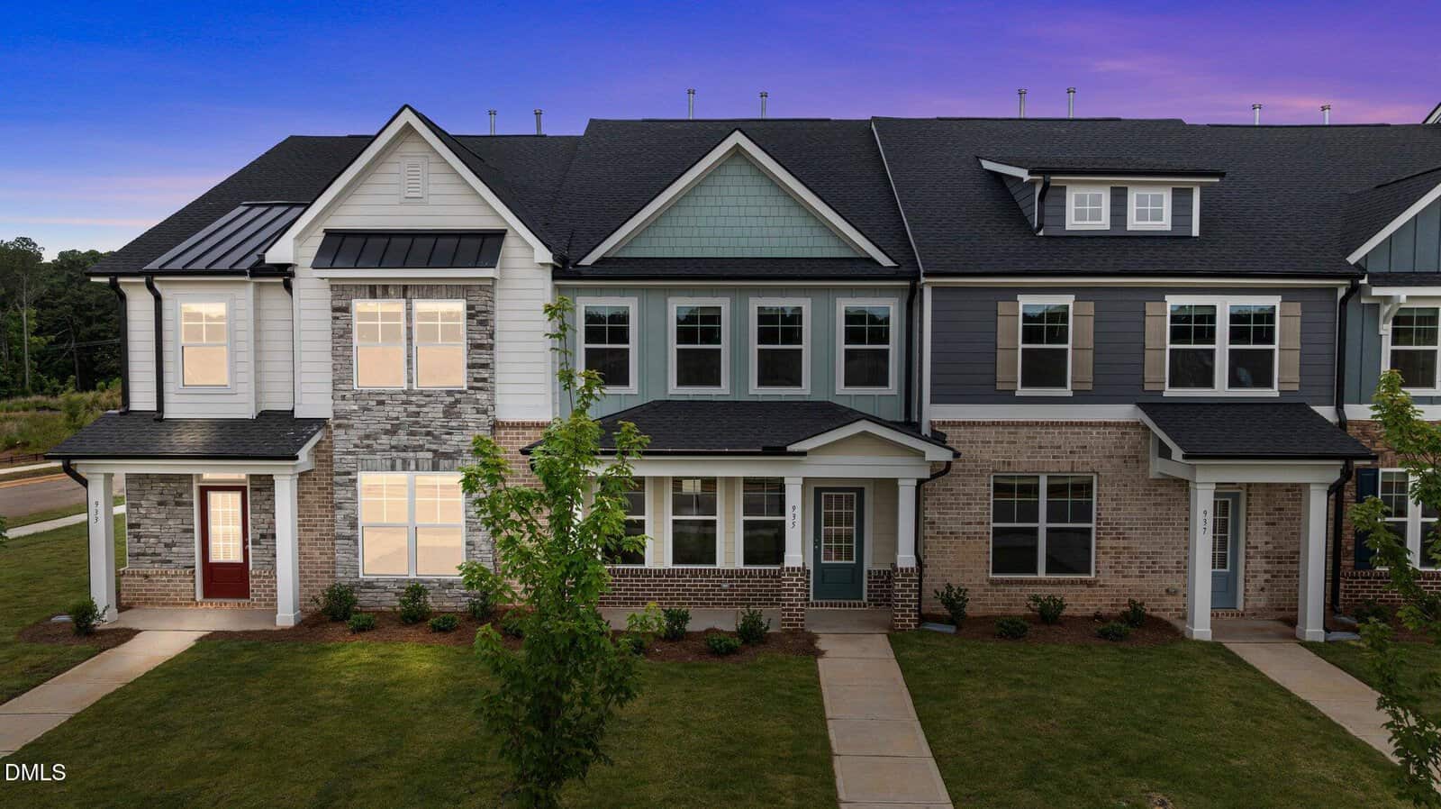 Two-story townhouse buildings in Wake Forest at 933 Alma Railway Drive feature brick, siding, and stone exteriors, covered front porches, sidewalk paths, and newly planted trees and shrubs, beautifully photographed at dusk with a purple sky.