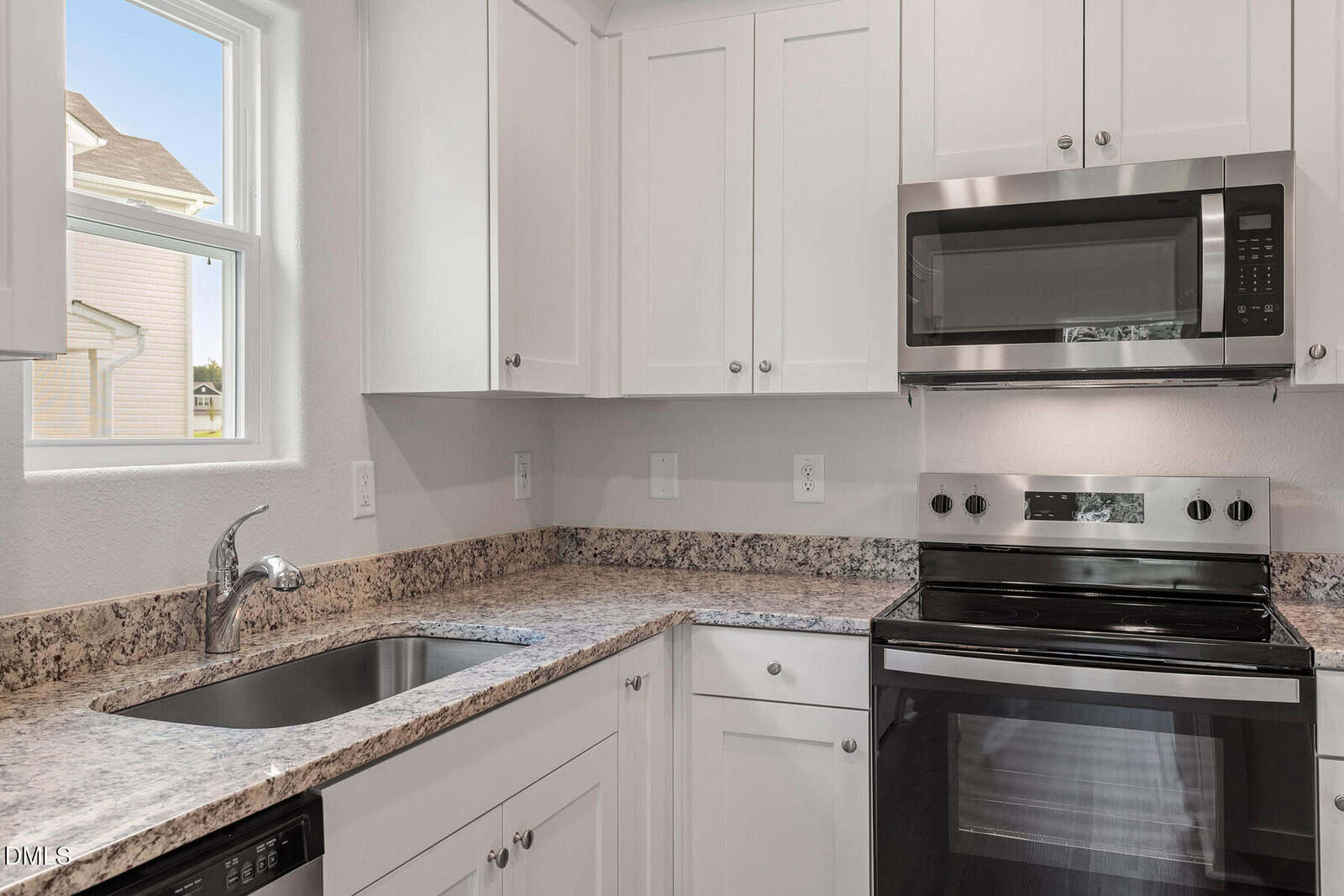 A modern kitchen with white cabinets, a built-in microwave above a stainless steel electric stove, granite countertops, a sink with a faucet under a window, and neutral-colored walls.