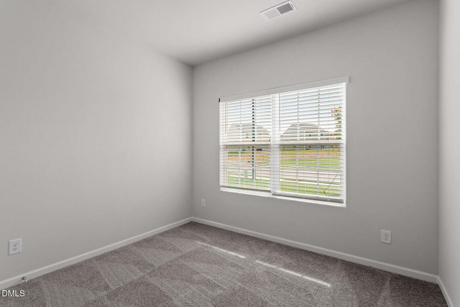 Empty room with light gray carpet, white walls, and a window with white blinds letting in natural light. The window shows a green lawn and part of a fence outside.