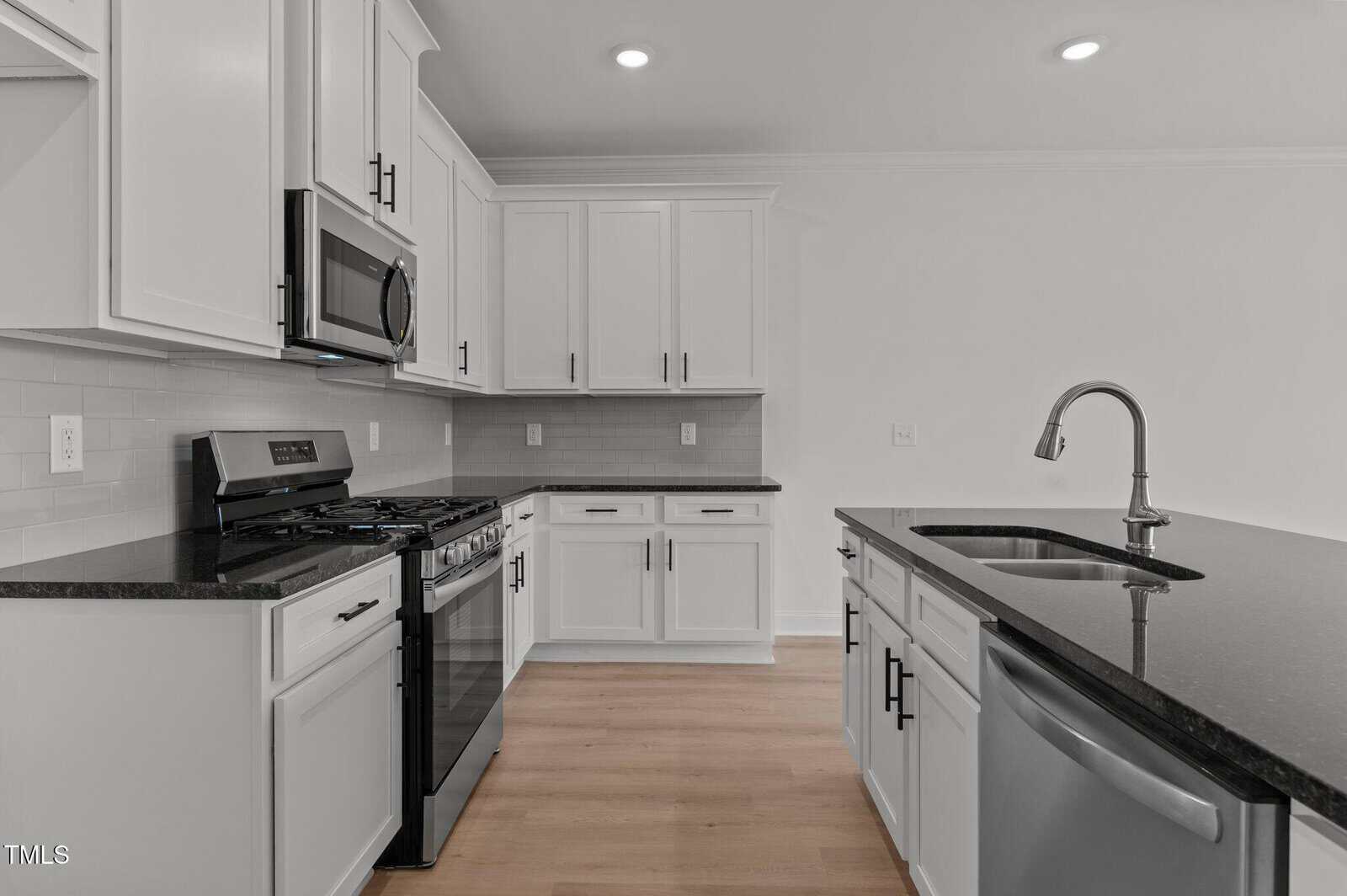 Modern kitchen with white cabinets, black countertops, stainless steel appliances including a gas stove, microwave, and dishwasher, an island with a sink, light wood flooring, and neutral-colored walls.