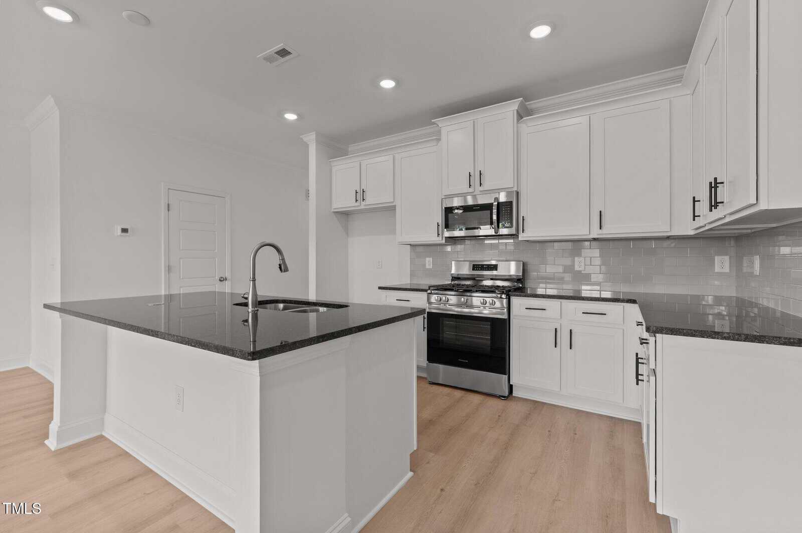 Modern kitchen with white cabinets, stainless steel appliances, a gas stove, built-in microwave, black countertops, a tile backsplash, and a center island with a sink. The floor is light wood, and the space is well-lit.