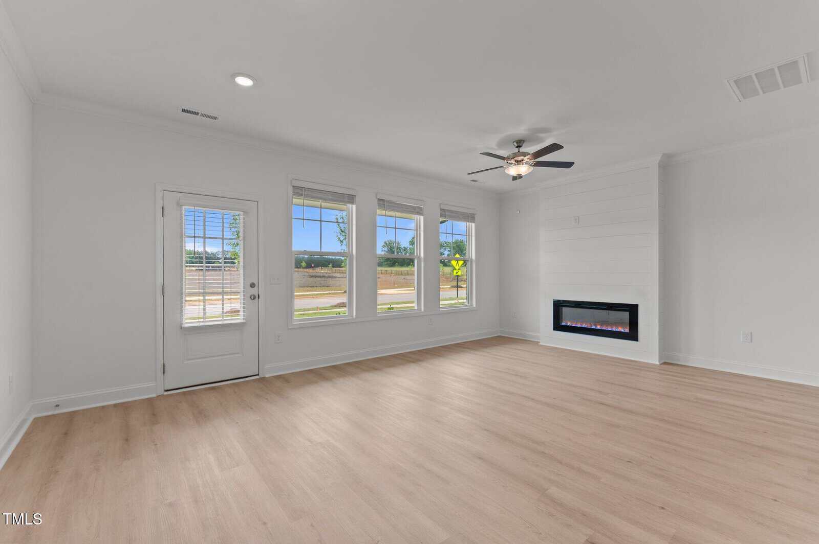 Bright, empty living room with light wood flooring, white walls, large windows, a glass door, ceiling fan, and a modern built-in electric fireplace on the right wall.