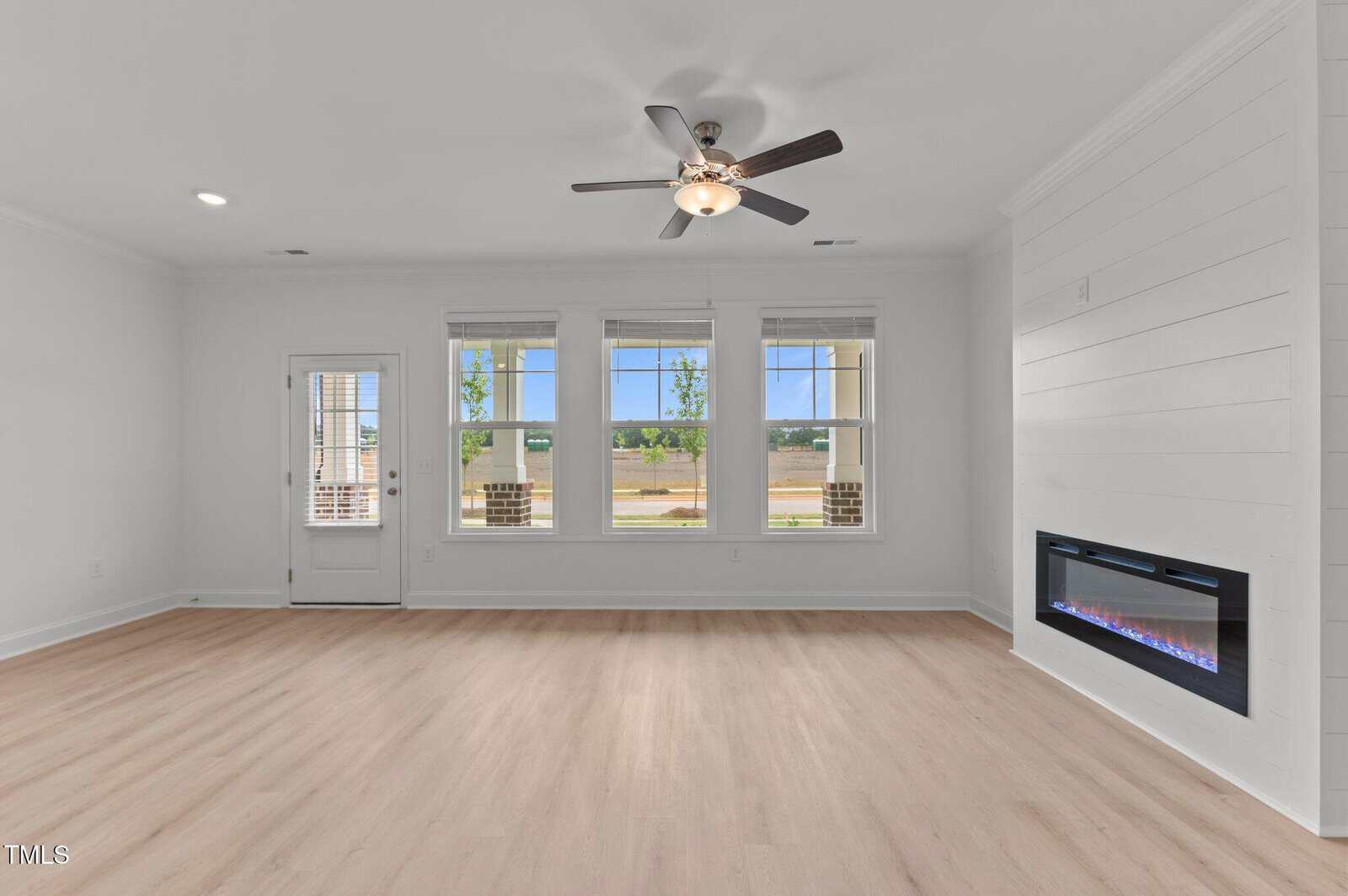 A bright, empty living room with light wood floors, white walls, large windows, a ceiling fan, a modern electric fireplace, and a door leading outside.