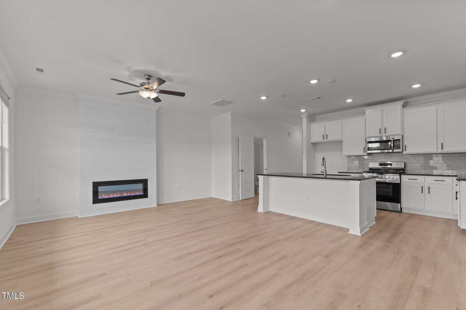 Modern open-concept living area with light wood floors, a ceiling fan, wall-mounted electric fireplace, and a kitchen featuring white cabinets, stainless steel appliances, and a large island with a dark countertop.