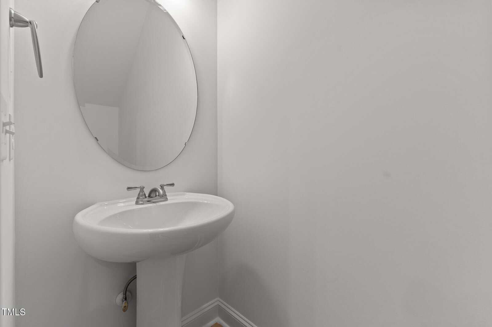 A small, minimalist bathroom with a white pedestal sink, a silver faucet, and an oval mirror mounted above the sink against plain white walls.