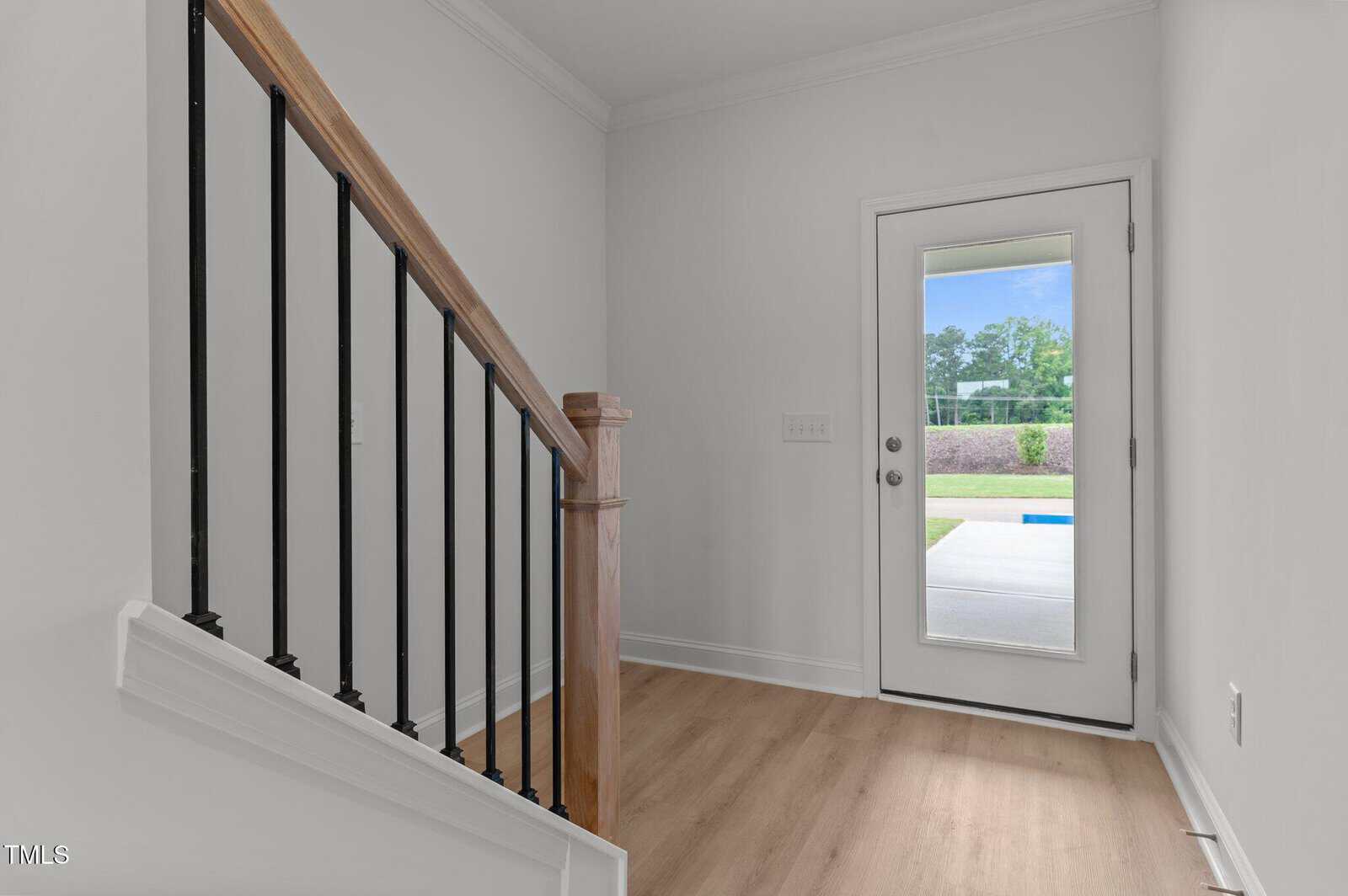 A bright entryway with light wood flooring, a staircase with black balusters and a wooden handrail, and a glass door leading outside to a patio and green yard.