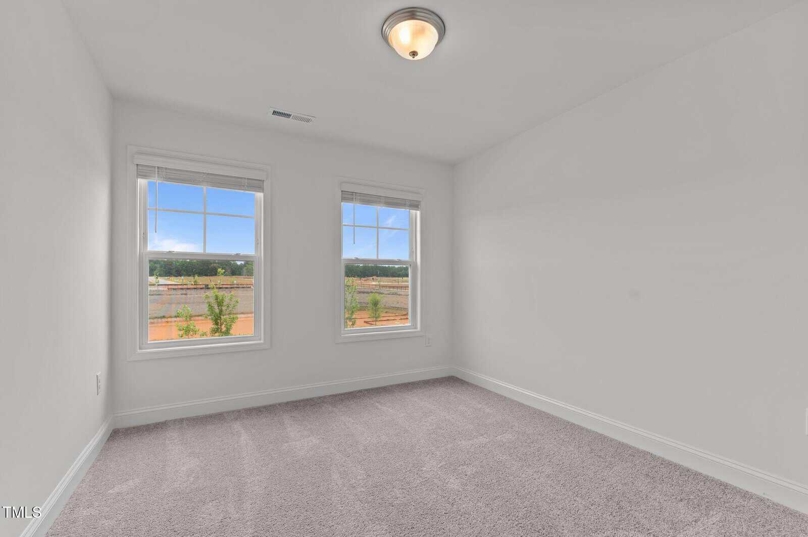 Empty room with light gray carpet, white walls, and two large windows showing an outdoor view with trees and blue sky. Ceiling has a single light fixture. Room is well-lit with natural sunlight.