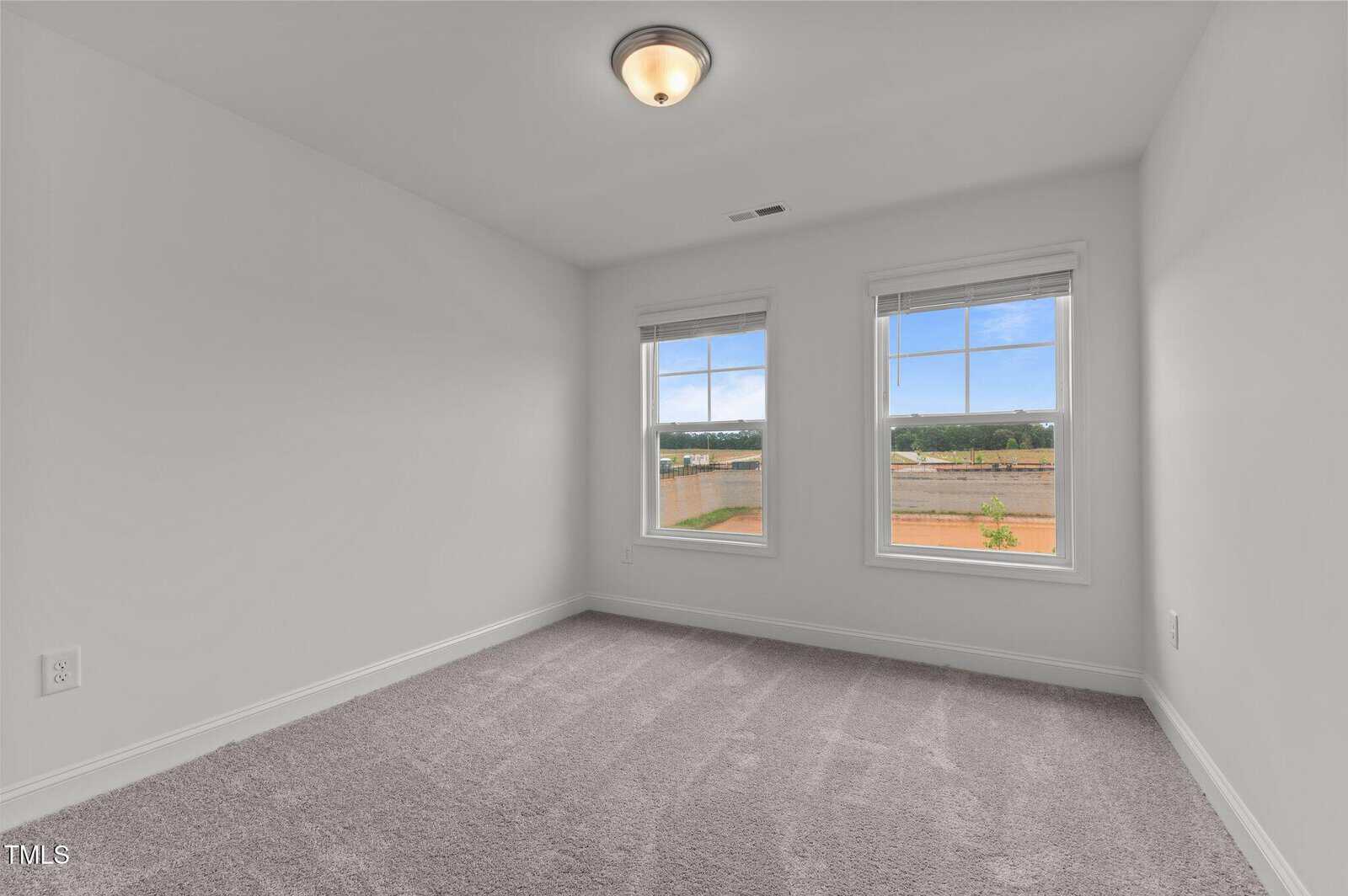 Empty, carpeted room with white walls, ceiling light fixture, two windows showing an outdoor view with blue sky, and an electrical outlet visible on the left wall.