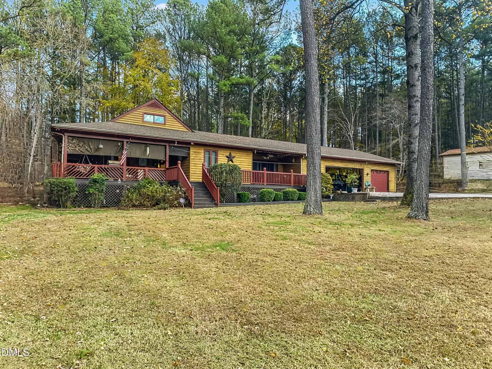 A long, single-story wooden house with a covered porch sits on a grassy lawn, surrounded by tall trees and a wooded area in the background. The house features red and brown trim and a two-car garage.