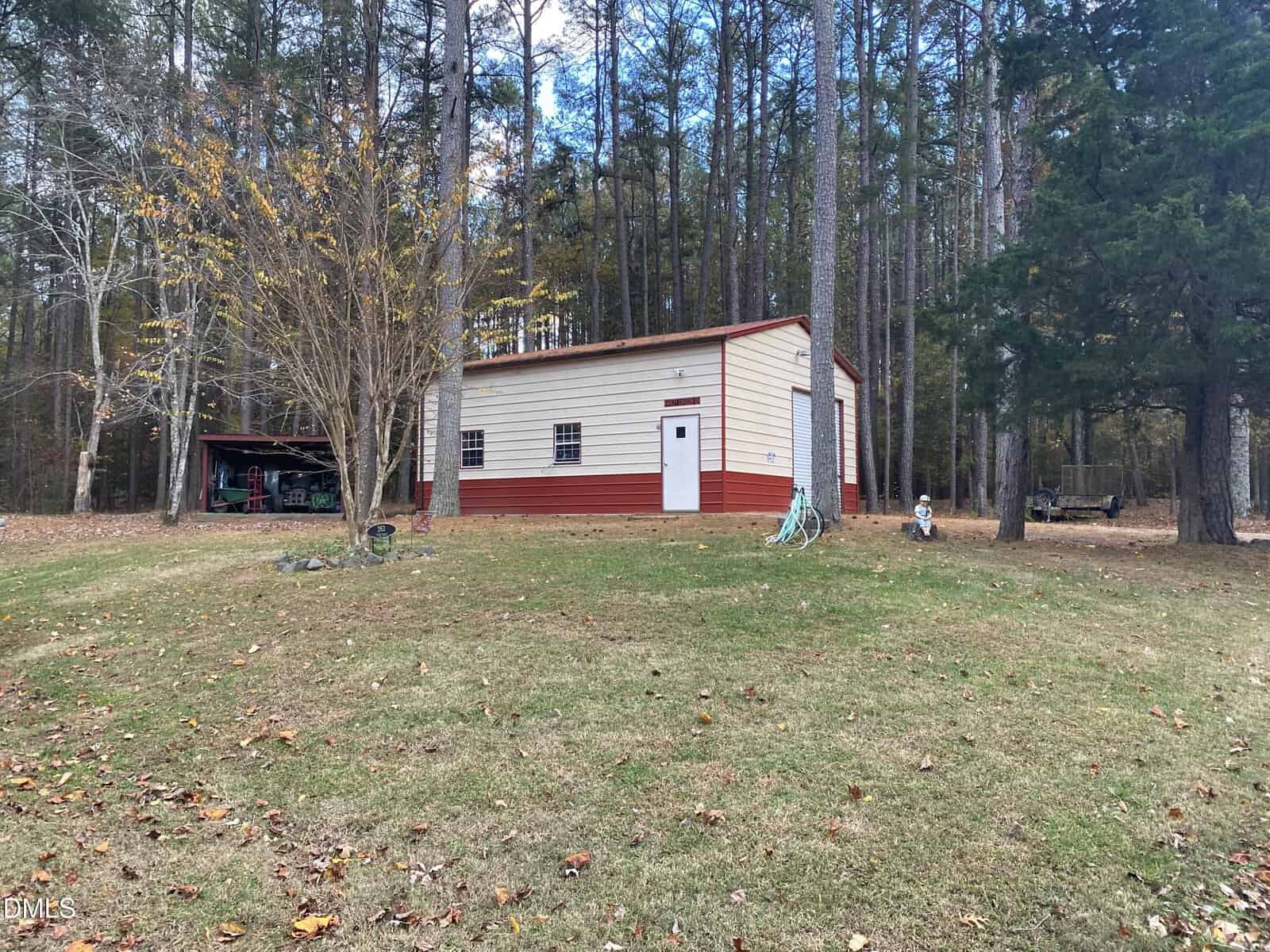 A small beige and red building with a metal roof sits in a grassy yard surrounded by tall trees. A hose is coiled on the ground, and a covered area with tools and items is visible to the left.