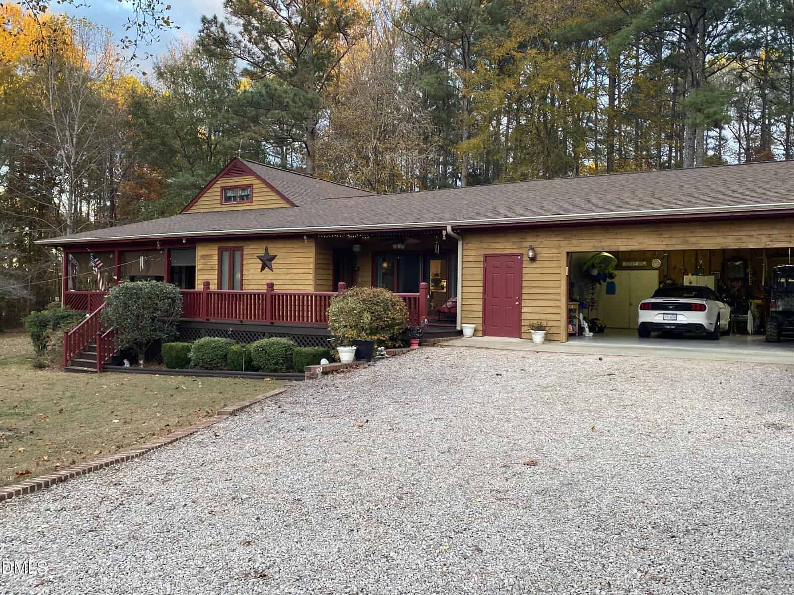 A one-story wooden house with a red porch, star decoration, and attached open garage, surrounded by trees with autumn foliage. A white car is parked inside the garage, and a gravel driveway is in front.