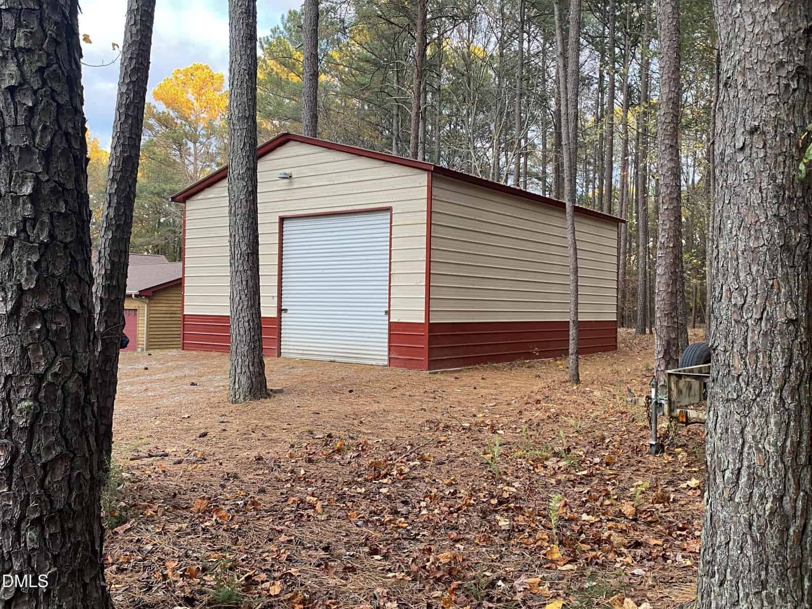A large beige and red metal storage building with a closed roll-up garage door sits among tall pine trees in a wooded area, with dry leaves and pine needles covering the ground.