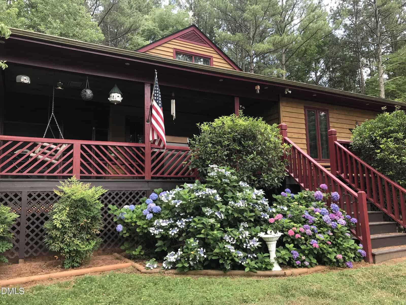 A charming wooden house at 263 Pine Trail with a covered front porch, red railings, an American flag, and blooming hydrangea bushes in the yard, surrounded by green trees and shrubs in Henderson.