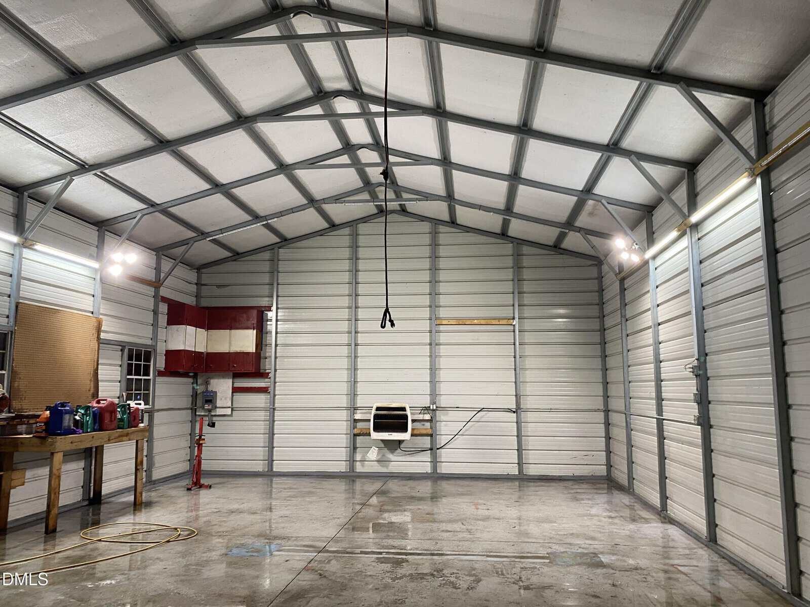 Interior of a clean, empty metal garage with a high ceiling, overhead lights, a workbench with tools on the left, red cabinets on the wall, and concrete floor with a rope hanging from the center beam.
