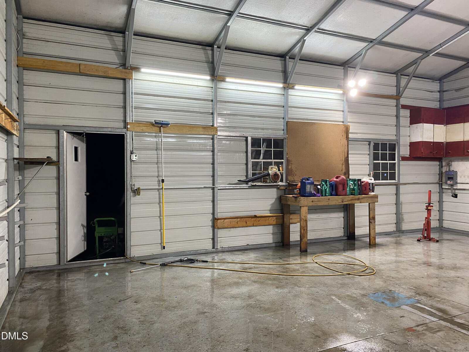 Interior of a garage with a wet floor, a broom on the wall, a workbench with colored fuel cans, tools, a coiled hose, two windows, an open door, and bright overhead lights.