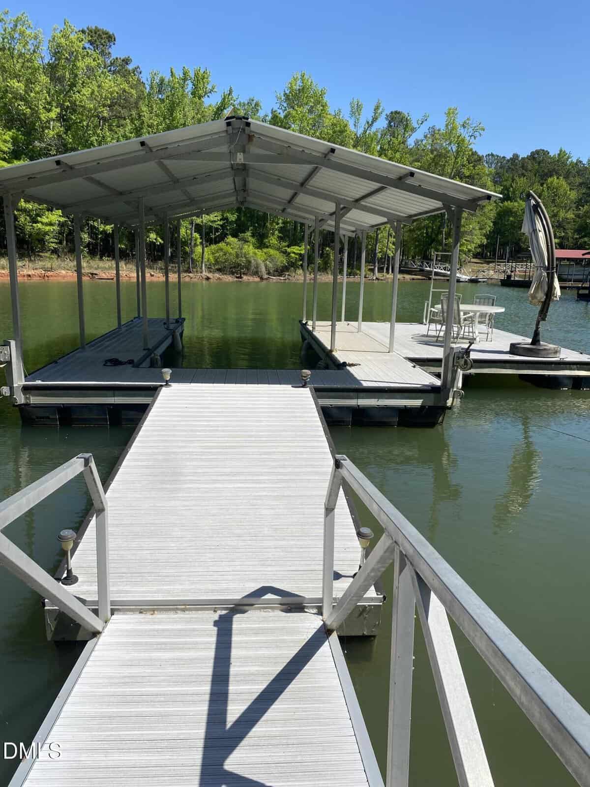 A covered boat dock with a walkway extends over a calm lake. There is a table and chairs on the dock to the right, surrounded by green trees under a clear blue sky.